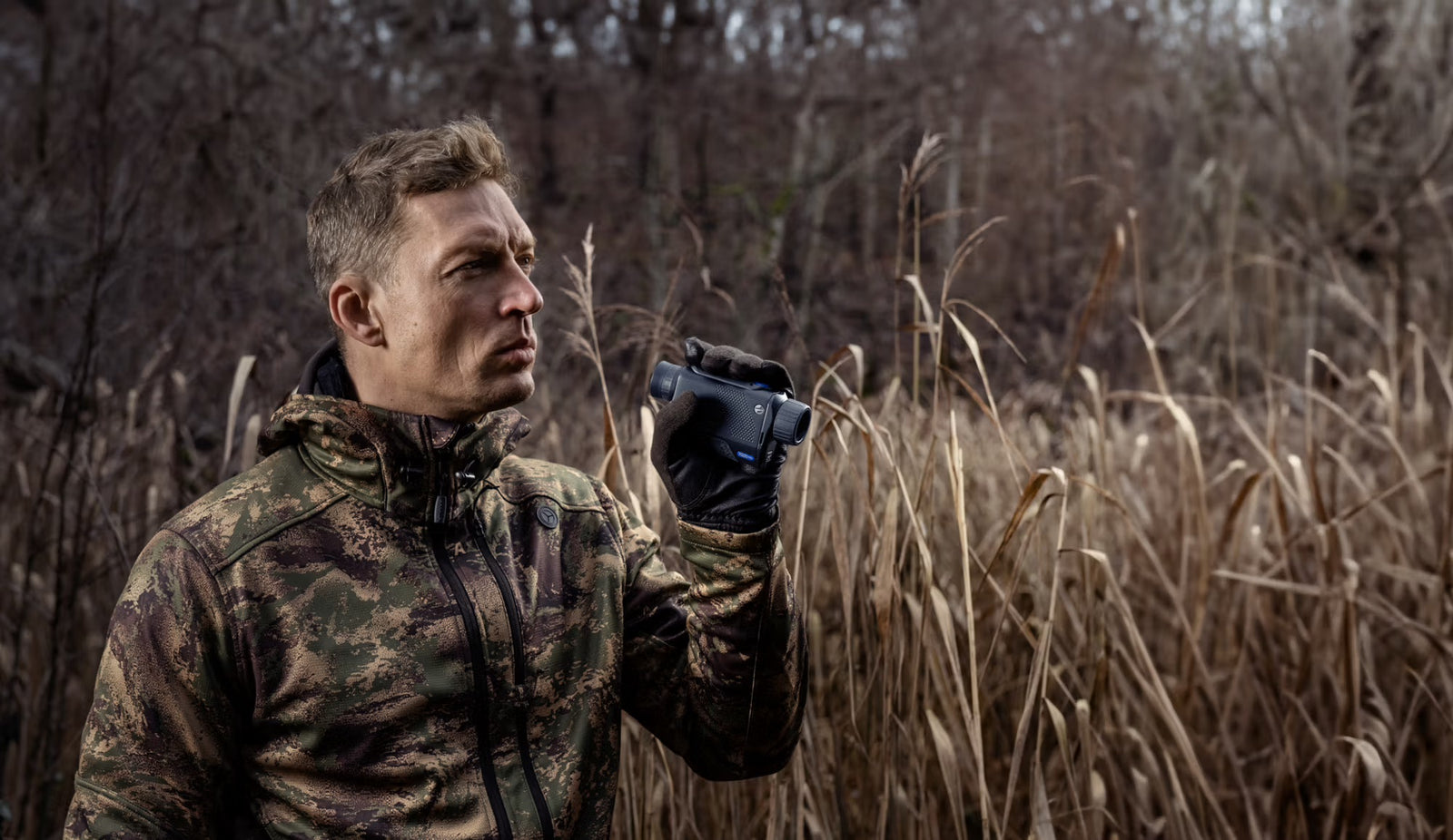 Ein Mann in Tarnjacke und schwarzen Handschuhen hält ein PULSAR Wärmebildgerät Axion XQ30 PRO in der Hand und scannt in der Nähe von hohem, trockenem Schilf und kahlen Bäumen, konzentriert auf seine Jagd.