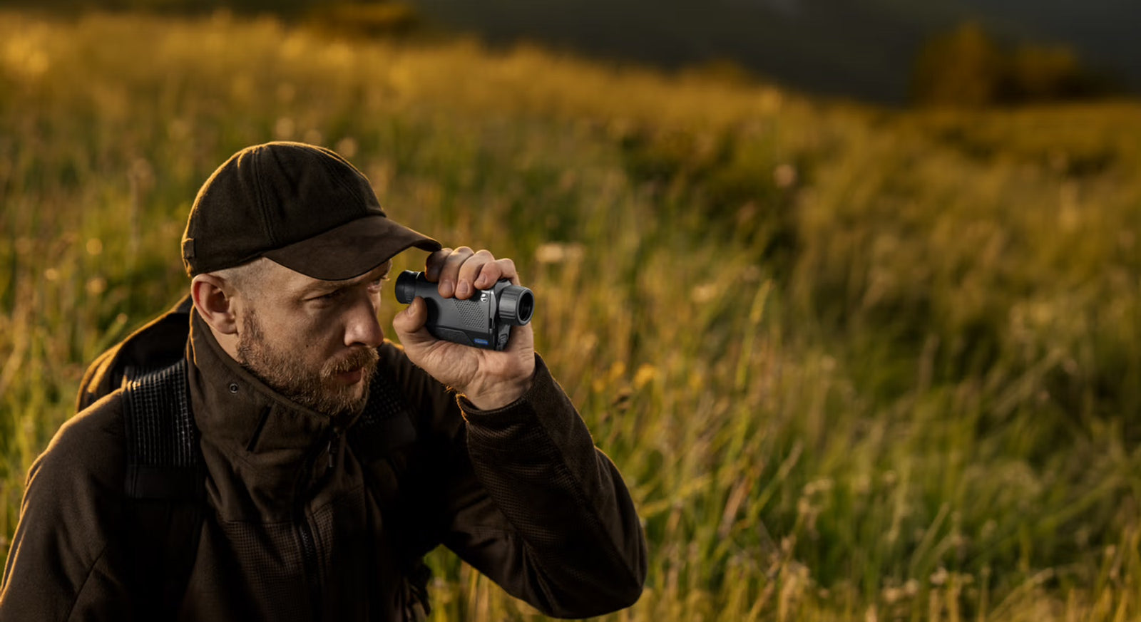 Ein Mann in Outdoor-Kleidung und Mütze benutzt ein PULSAR Pulsar Wärmebildgerät Axion XQ30 PRO, während er bei Sonnenuntergang auf einer Wiese steht und sich auf die Jagd vorbereitet, mit unscharfen Hügeln im Hintergrund.