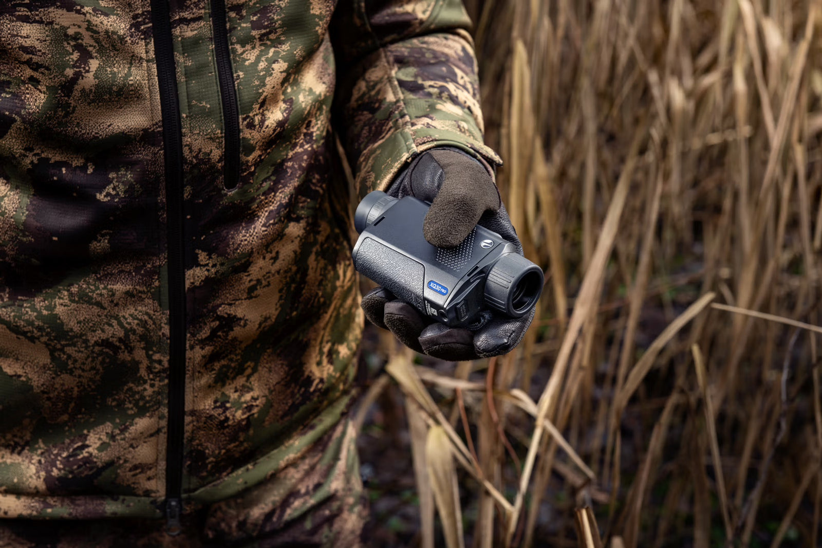 Eine Person in Camouflage hält ein PULSAR Wärmebildgerät Axion XQ30 PRO, das sich perfekt für die Jagd eignet, in einer grasbewachsenen Umgebung.