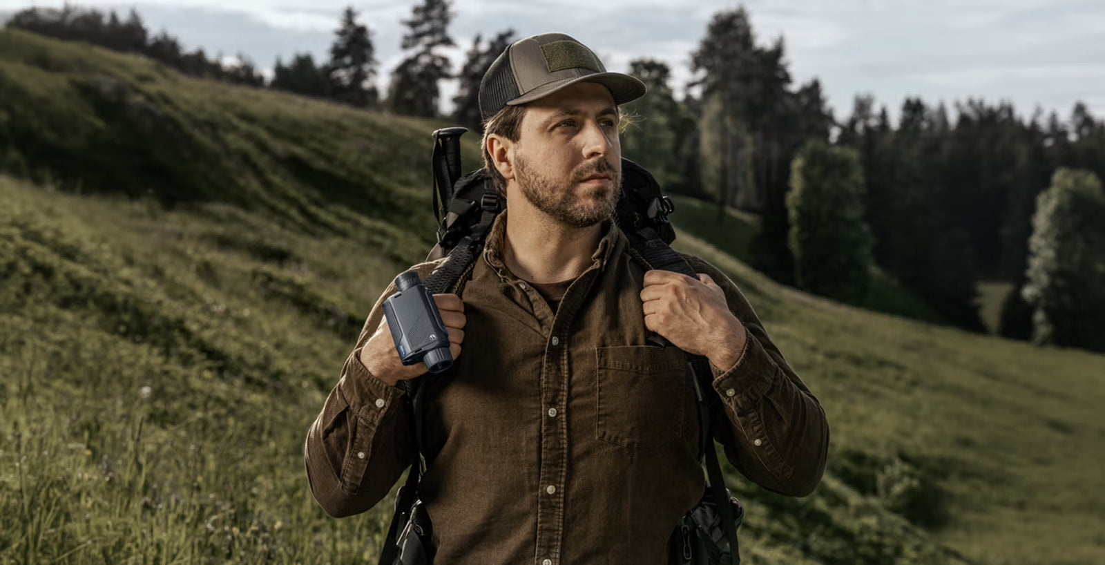 Ein Mann in braunem Hemd und Mütze steht auf einer grasbewachsenen Anhöhe, hält seinen Rucksack und blickt mit dem PULSAR Wärmebildgerät Axion XQ30 PRO - ideal für die Jagd - zwischen Bäumen und bewölktem Himmel nach vorne.
