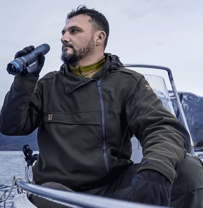 Ein Mann in grüner Jacke sitzt in einem Boot auf einem See, hält das PULSAR Wärmebildgerät Telos LRF XP50 und blickt in die Ferne. Im Hintergrund sind schneebedeckte Berge und ein bewölkter Himmel zu sehen.