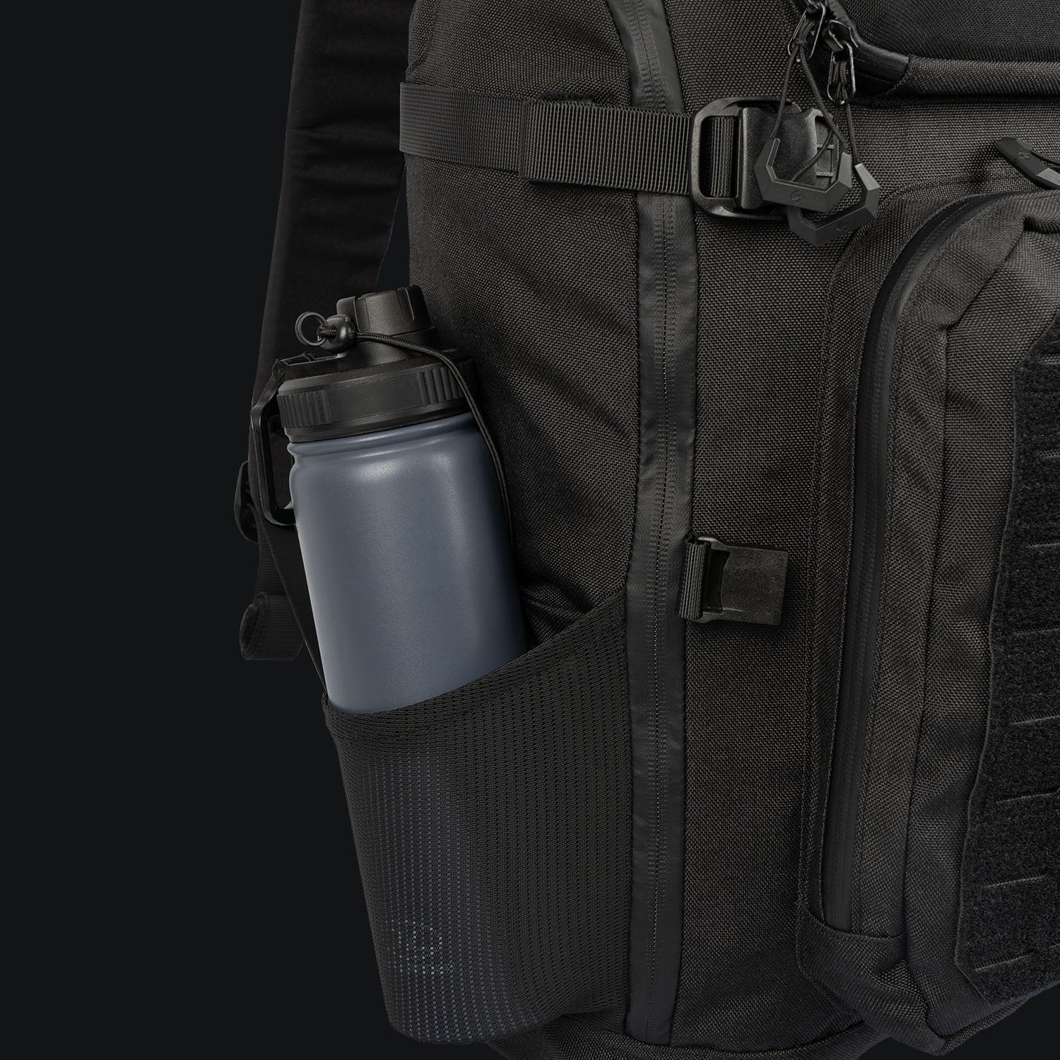 Eine Nahaufnahme des STOIRM 25L Tactical Pack GEN2 von STOIRM zeigt einen schwarzen Rucksack mit einer grauen Wasserflasche in der seitlichen Netztasche, mit Reißverschlussdetails und verstellbaren Trägern vor einem dunklen Hintergrund.