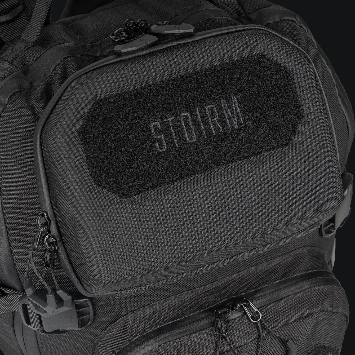 Nahaufnahme des STOIRM 25L Tactical Pack GEN2 in Schwarz, mit mehreren Reißverschlüssen und Fächern. Auf der Vordertasche befindet sich ein STOIRM-Aufnäher in hellgrauen Großbuchstaben. Perfekt für den Transport Ihrer Outdoor-Ausrüstung.