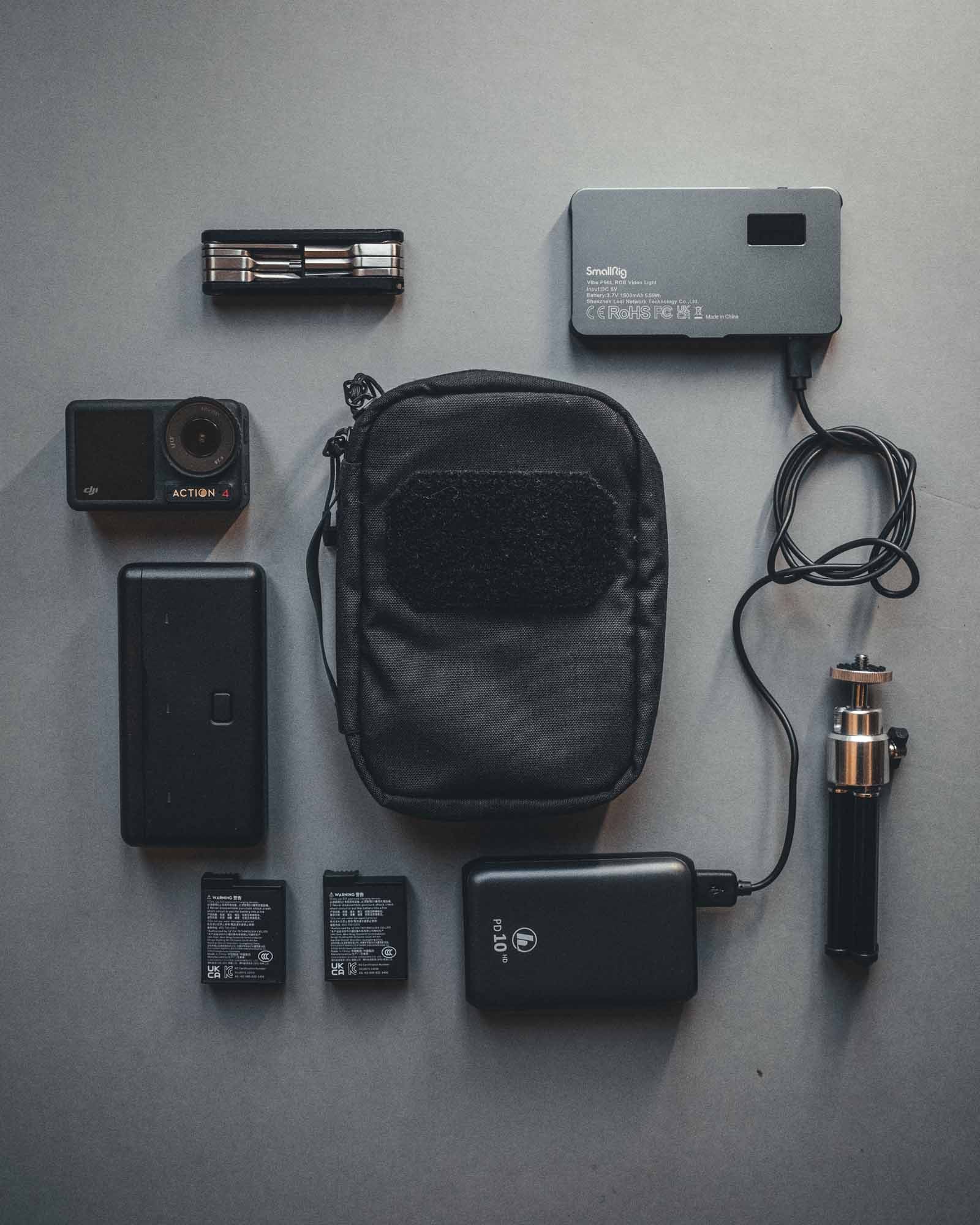 Eine flache Anordnung von technischer Ausrüstung - Kompaktkamera, Akkus, Ladegerät mit Kabel, Mini-Stativ, Powerbank - auf einer grauen Oberfläche mit der olivgrünen STOIRM Large Pouch V.1 von STOIRM zur organisierten Aufbewahrung.