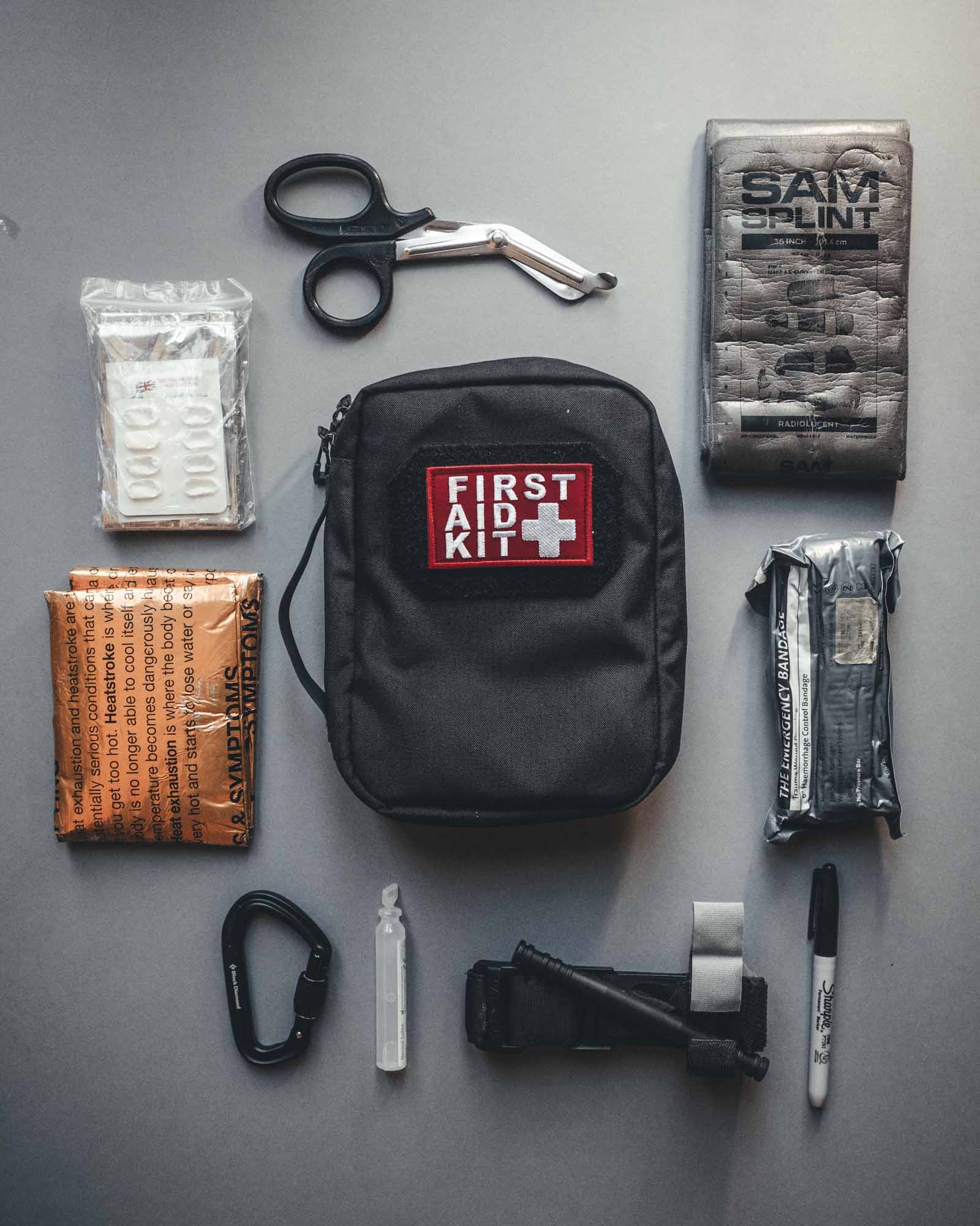 Auf grauem Hintergrund ist ein ordentlich organisiertes STOIRM Large Pouch V.2 Erste-Hilfe-Set abgebildet, das eine medizinische Schere, Verbandszeug, Handschuhe, einen Sharpie-Marker, einen Karabiner, Mull, einen Druckverband, ein Pillenpaket und mehr enthält.