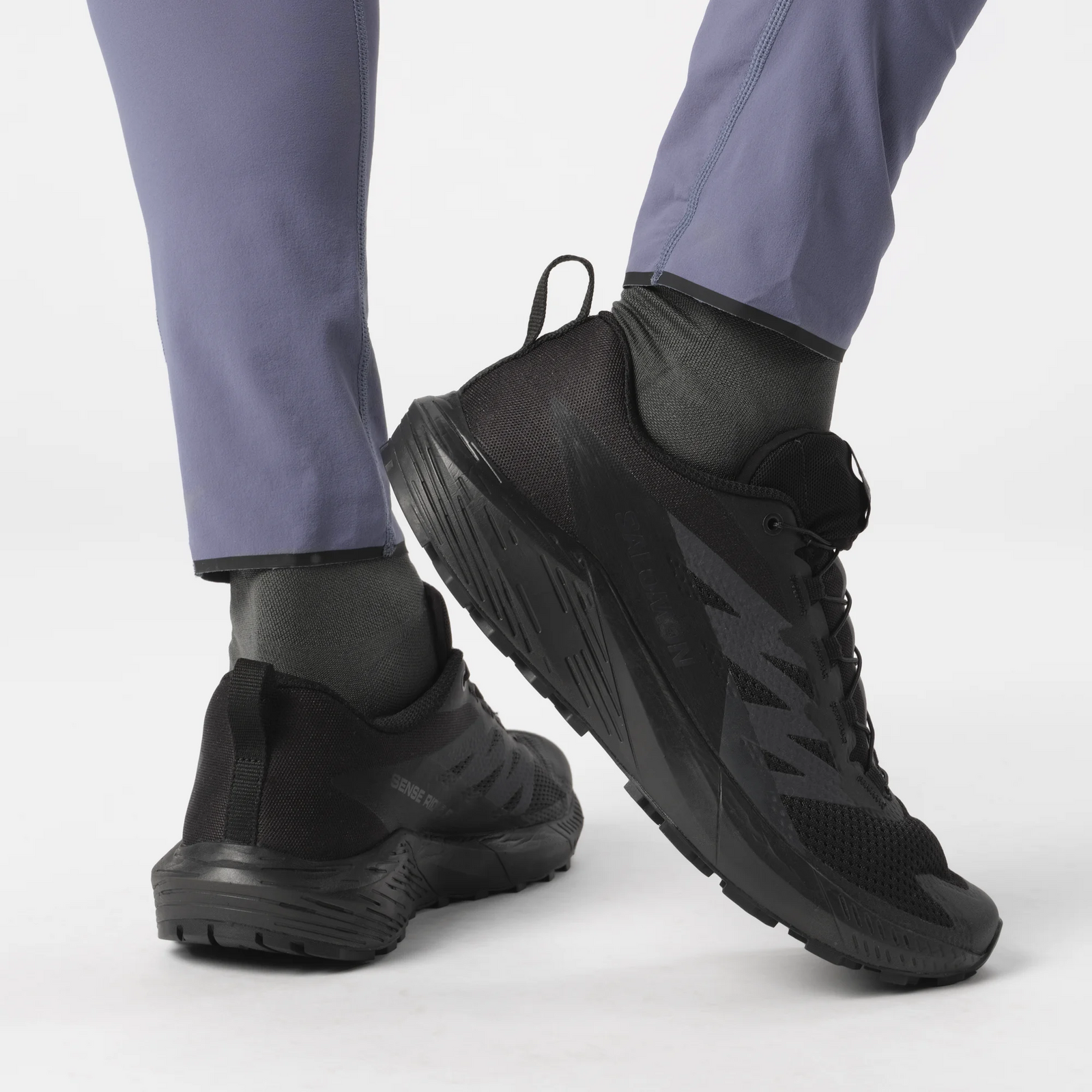 Nahaufnahme einer Person, die schwarze SALOMON FORCES Salomon SENSE RIDE 5 SR Trail-Schuhe mit All Terrain Contagrip-Sohle und eine schieferblaue Sporthose trägt, die von den Knien abwärts vor einem weißen Hintergrund gezeigt wird.