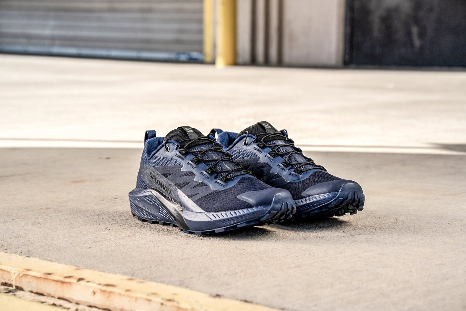 Ein Paar SALOMON FORCES Salomon SENSE RIDE 5 SR - India Ink Schuhe mit robusten All Terrain Contagrip Sohlen sitzen auf Beton neben einem gelben Bordstein.