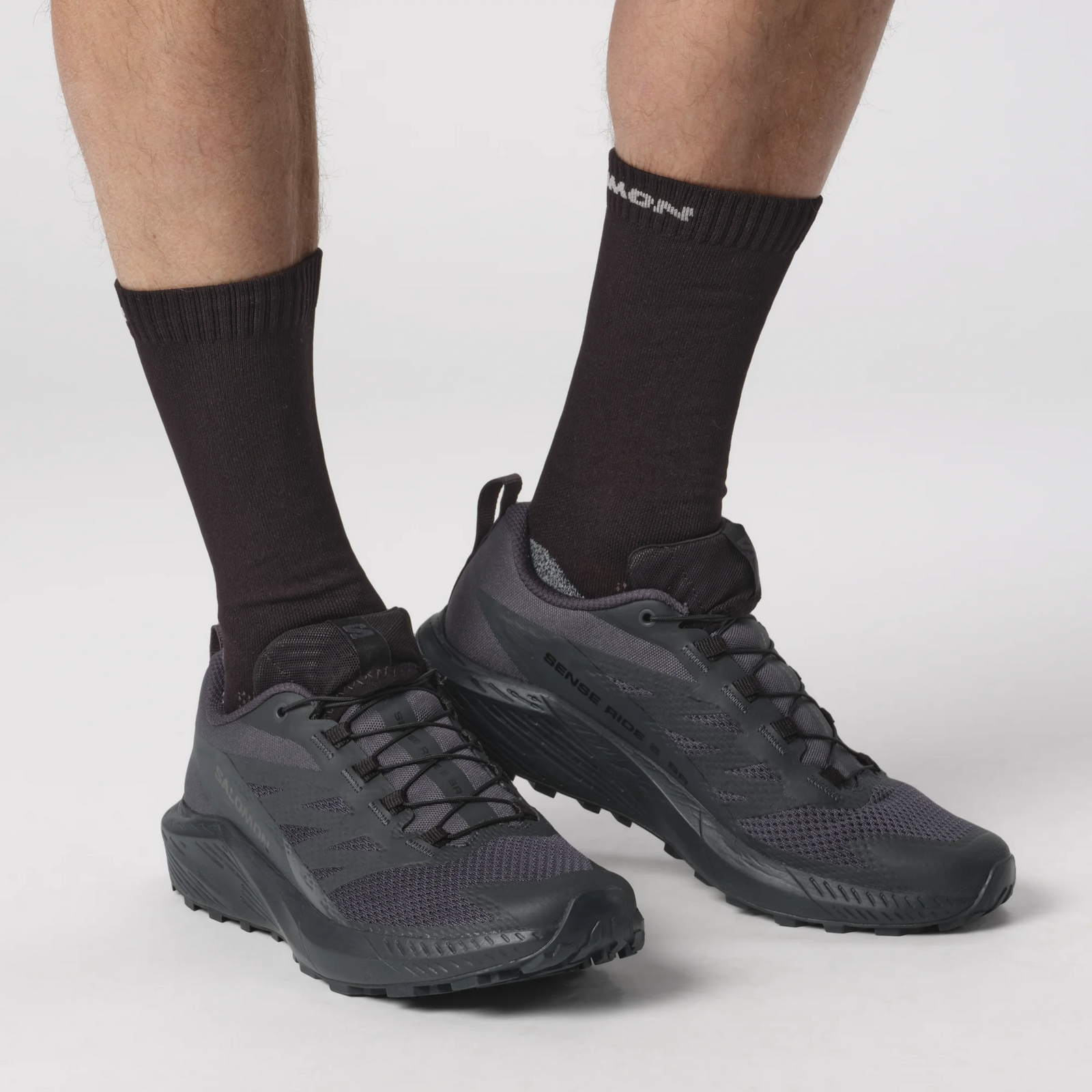Nahaufnahme einer Person, die SALOMON FORCES Salomon SENSE RIDE 5 SR - India Ink Trail-Schuhe mit All Terrain Contagrip-Sohle und schwarzen Socken trägt und nur ihre Unterschenkel und Füße vor einem weißen Hintergrund zeigt.