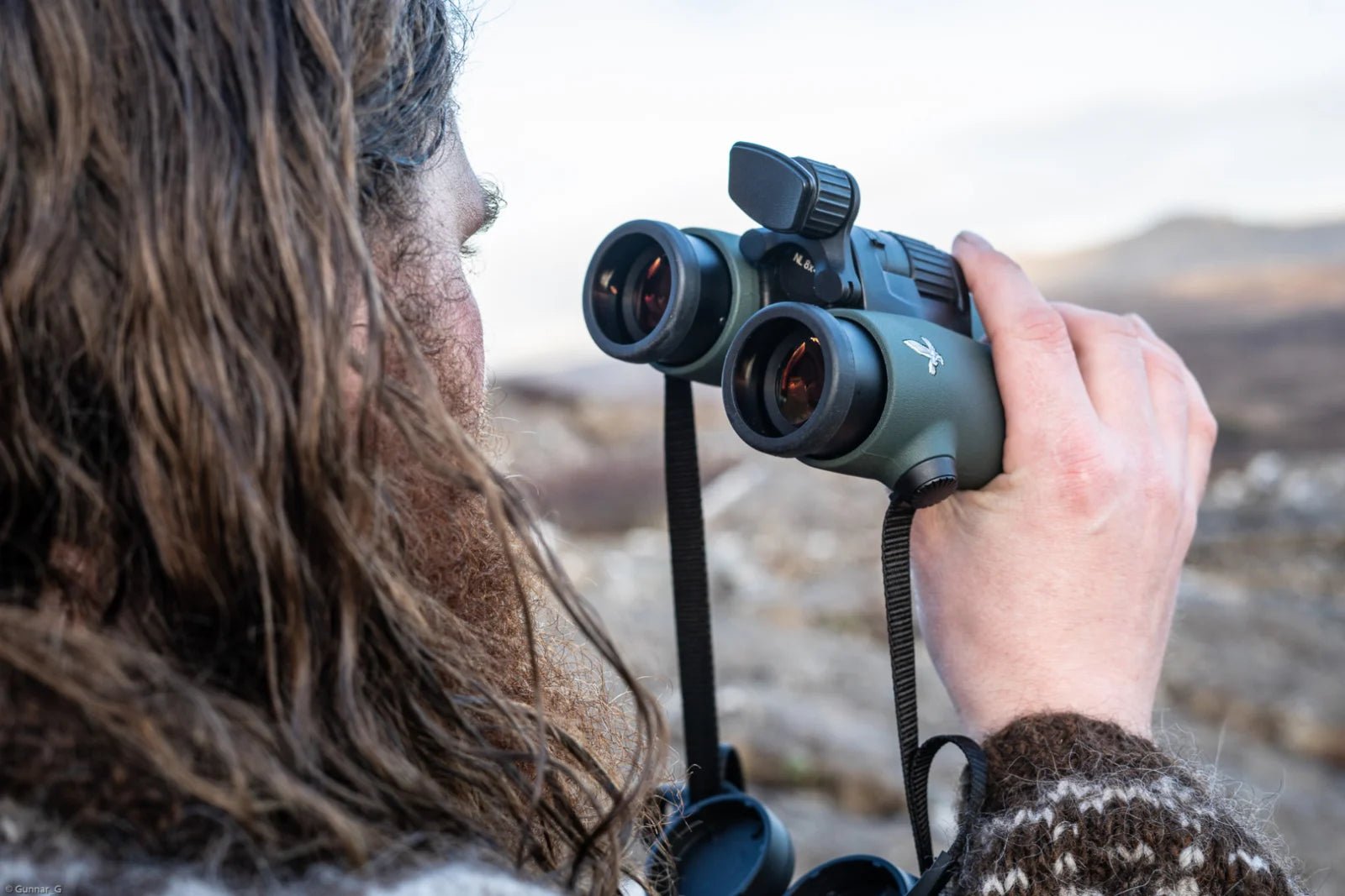 Eine Person mit langen braunen Haaren und einem gemusterten Pullover tastet mit der SWAROVSKI OPTIK FRP Stirnstütze NL Pure eine felsige Outdoor-Szene ab und zeigt dabei hochwertiges Fernglas-Zubehör für Outdoor-Fans.