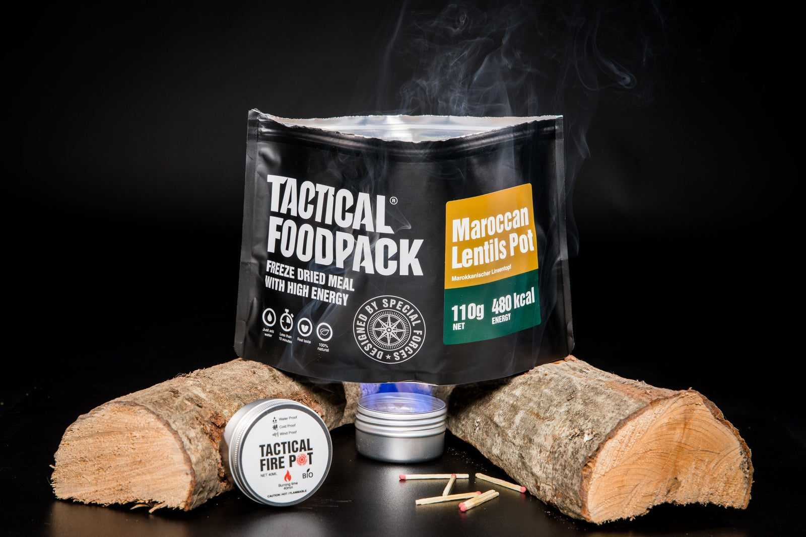 Ein TACTICAL FOODPACK Tactical Fire Pot glänzt vor einem gefriergetrockneten marokkanischen Linsentopf zwischen zwei Holzscheiten. Streichhölzer und eine brennende Flamme sorgen für Wärme, während Dampf aufsteigt, alles vor einem kräftigen schwarzen Hintergrund.