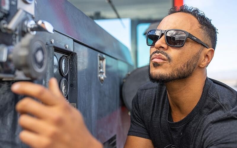 Ein Mann in schwarzem T-Shirt und WILEY X Wiley X Sonnenbrille TREK verstellt im Freien ein Bedienfeld und dreht konzentriert an einem Knopf der Maschine.