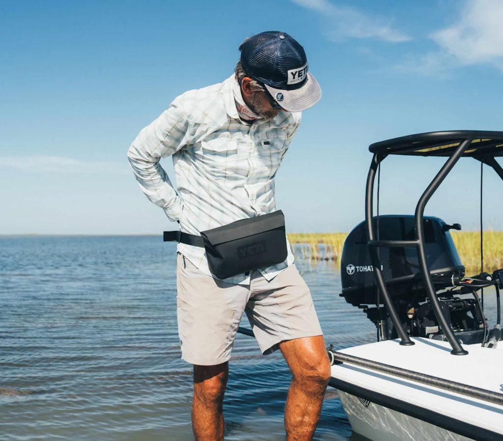 Ein Mann mit Mütze, Sonnenbrille, langärmeligem Hemd und Shorts passt seinen YETI® Sideclick® Gurt - Black an, während er an einem klaren Tag mit ruhigem Wasser im flachen Wasser neben einem Boot steht.