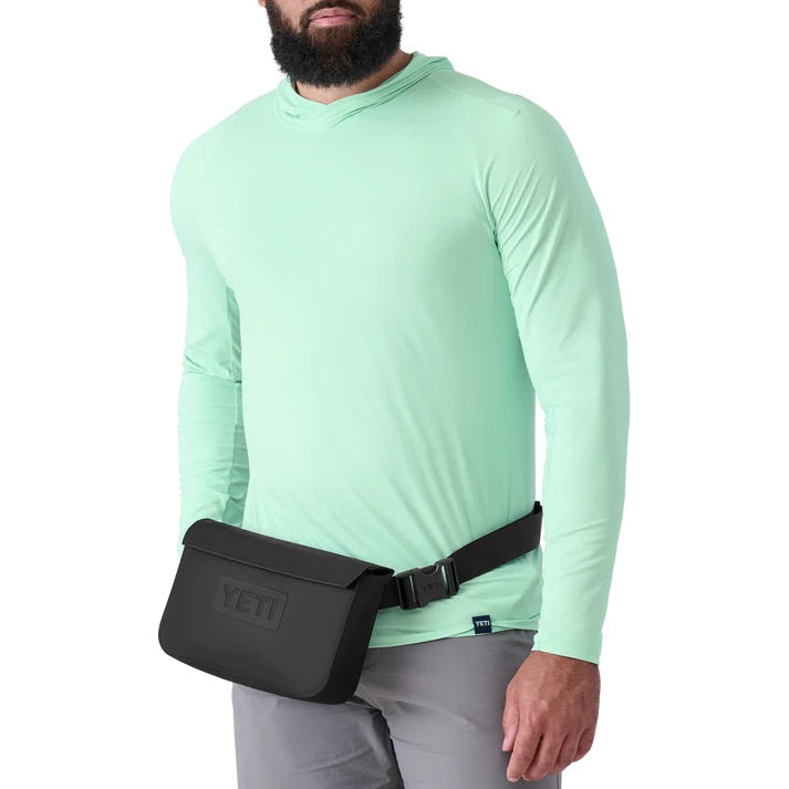Eine Person in einem hellgrünen langärmeligen Hemd und einer grauen Hose steht mit dem YETI® Sideclick® Gurt in Schwarz von YETI, der aus strapazierfähigem Nylon besteht und in der Länge verstellbar ist, um die Hüften. Der Kopf ist teilweise aus dem Bild.