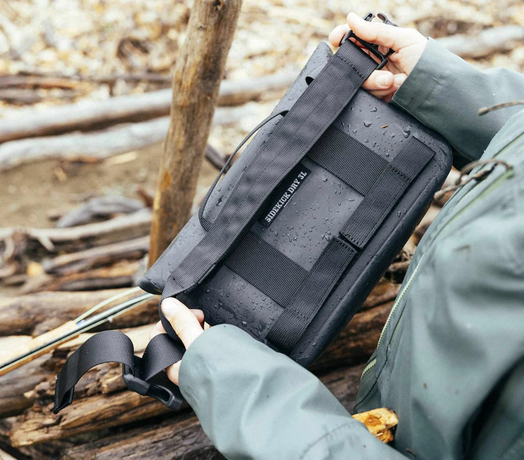 Eine Person in grüner Jacke hält den YETI® Sideclick® Gurt - Black, eine wasserfeste Tasche aus strapazierfähigem Nylon mit Regentropfen darauf, im Freien zwischen nassen Ästen und Baumstämmen stehend.