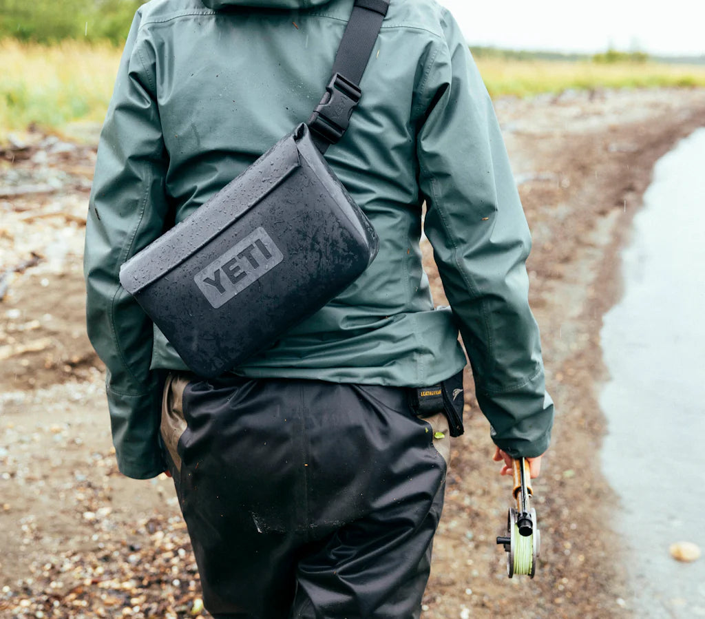 Eine Person in grüner Jacke und wasserdichter Hose geht an einem See spazieren, trägt eine Fliegenfischerrolle und einen schwarzen YETI® Sideclick® Gurt von YETI aus strapazierfähigem Nylon mit verstellbarer Länge.