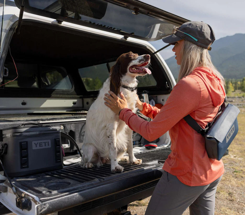 Eine Frau in Outdoor-Ausrüstung streichelt einen glücklichen Hund auf der offenen Heckklappe eines Pickups. In der Nähe stehen eine YETI-Kühlbox und ein schwarzer YETI® Sideclick® Gurt, während sich im Hintergrund schroffe Berge erheben.