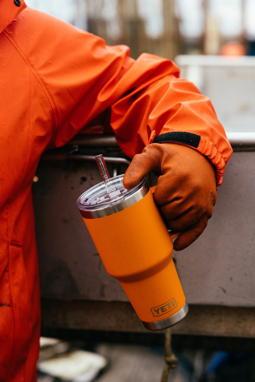 Eine Person in orangefarbener Jacke und Handschuhen hält einen YETI® Becher Rambler 35 Oz Straw Mug - King Crab Orange mit doppelwandiger Vakuumisolierung, während sie im Freien in der Nähe einer Metallfläche steht.