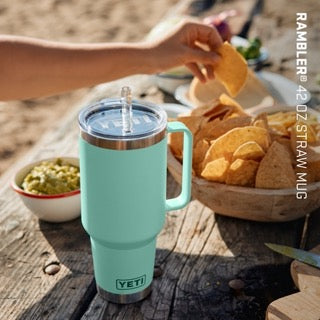 Ein YETI® Becher Rambler 42 Oz Straw Mug in Seafoam steht auf einem Picknicktisch neben Tortillachips und Guacamole, während draußen am Strand eine Hand nach einem Chip greift.