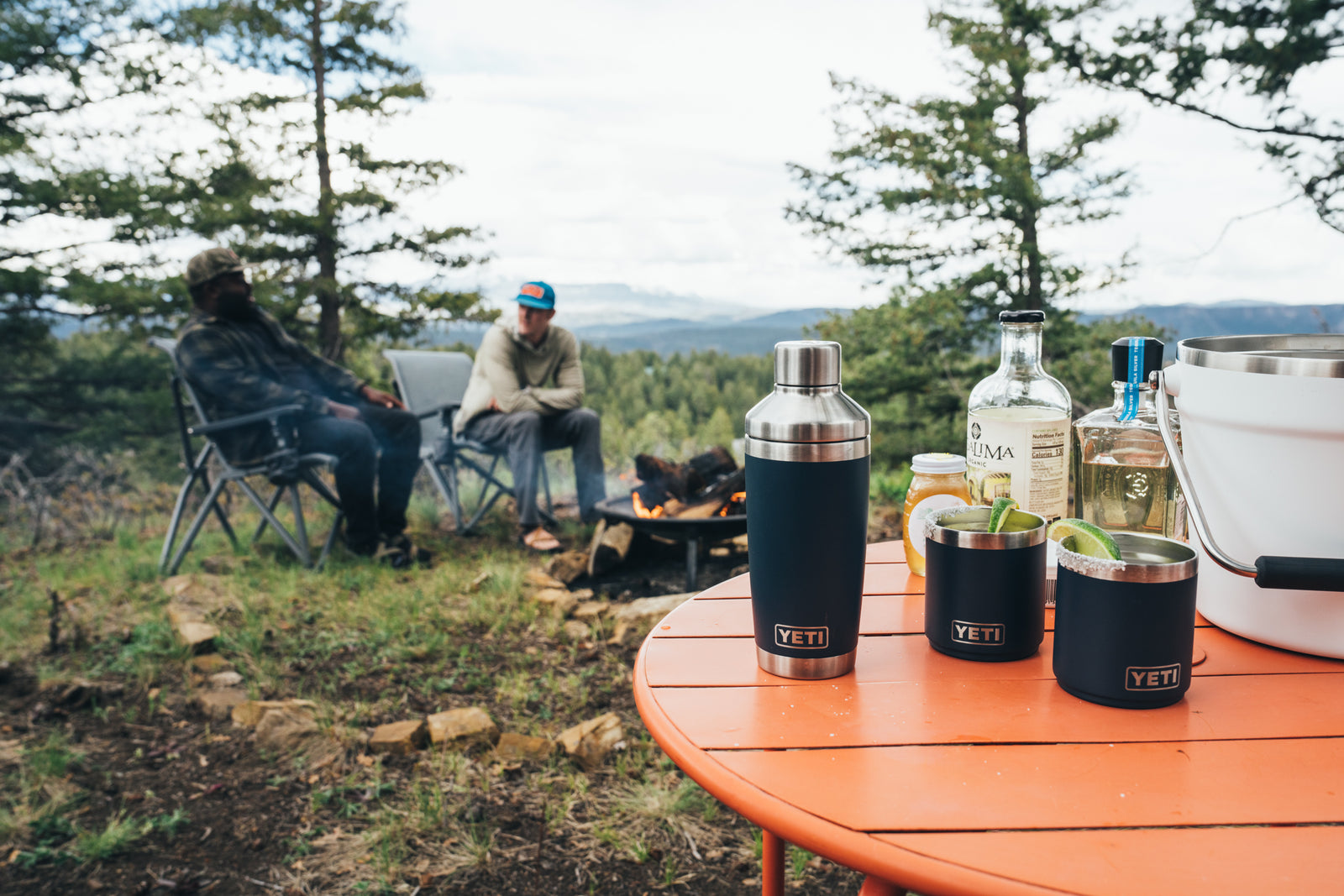 Zwei Personen sitzen an einem Lagerfeuer im Wald, während auf dem roten Tisch im Vordergrund Getränke, Flaschen und ein YETI® Cocktail-Shaker 20 Oz - Navy stehen. Bäume und Berge in der Ferne bilden eine friedliche Kulisse.