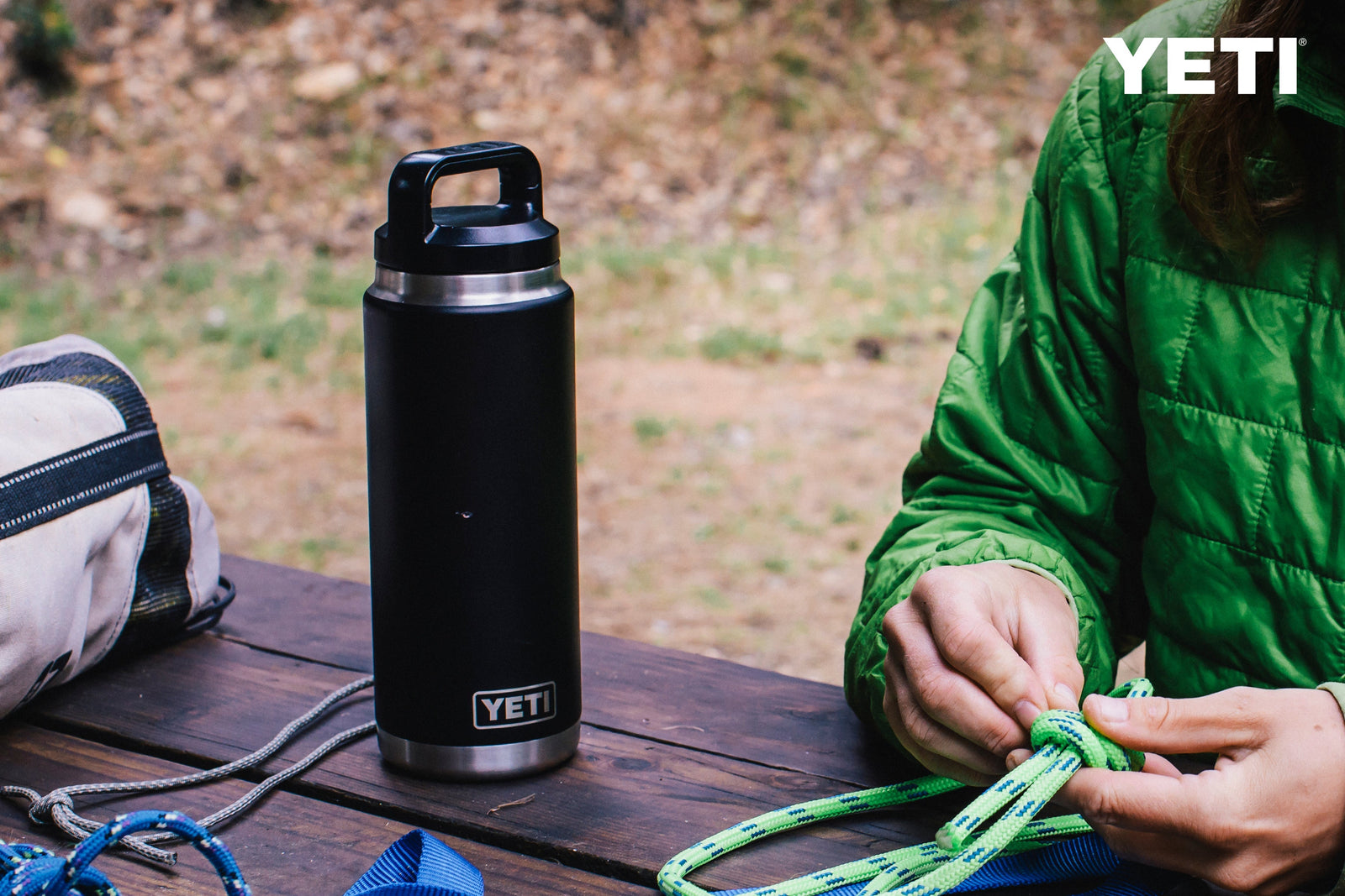 Eine YETI® Flasche Rambler 26 Oz - Navy steht auf einem Holztisch im Freien, während eine Person in grüner Jacke in der Nähe ein Kletterseil knüpft, dessen Ausrüstung sichtbar ist. Das YETI-Logo erscheint in der oberen rechten Ecke.