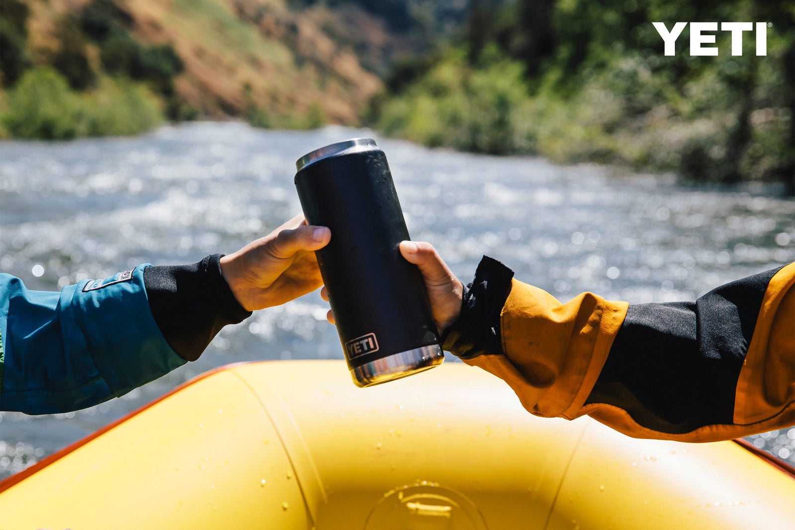 Zwei Hände reichen eine Navy YETI® Flasche Rambler 26 Oz mit auslaufsicherem Strohdeckel auf einem gelben Floß vor einem rauschenden Fluss und grünen Hügeln.