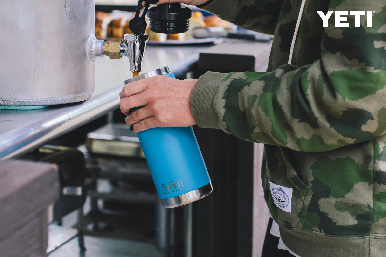 Jemand in einer Tarnjacke füllt eine YETI® Flasche Rambler 26 Oz - Navy mit Bier an einer Bar, deren auslaufsicherer Trinkhalmdeckel von Isolierflasche Edelstahl inspiriert ist.