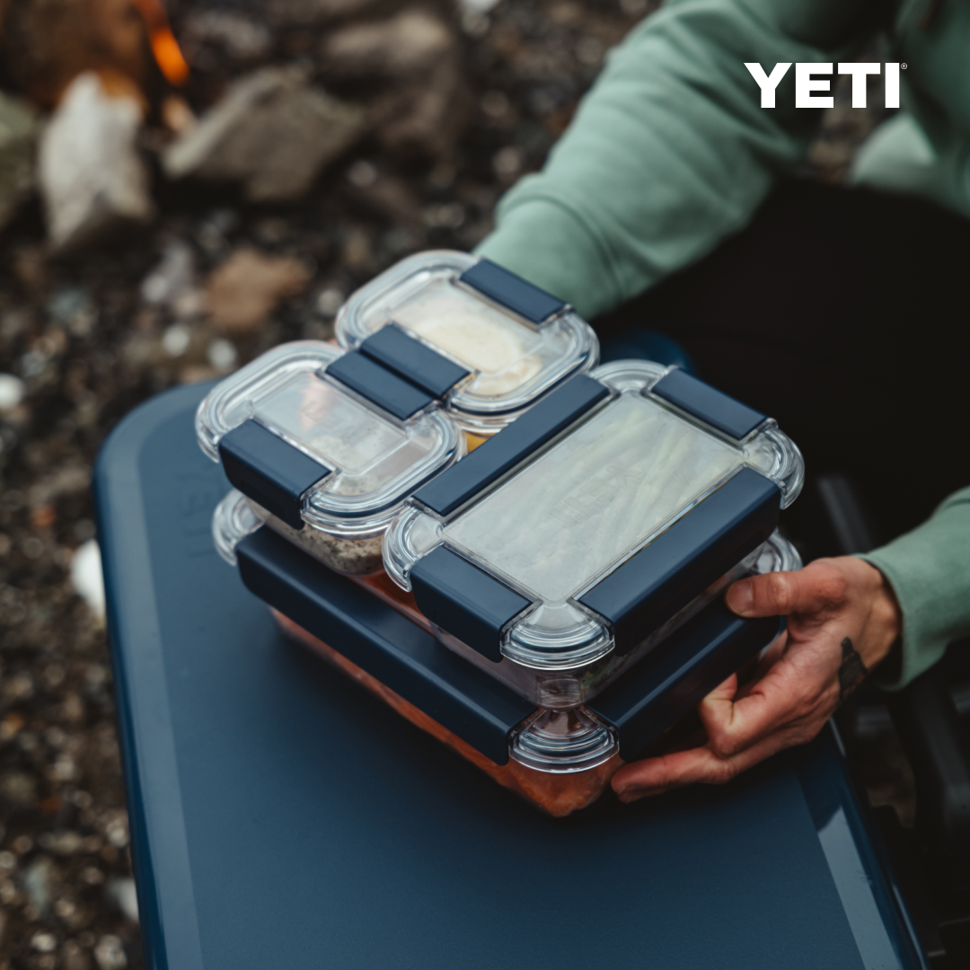 Eine Person hält einen Stapel von YETI® Food Storage Small-Behältern mit marineblauen Deckeln, die im Freien auf einer blauen Fläche stehen. Das YETI-Logo erscheint oben rechts, während im Hintergrund Felsen und ein Lagerfeuer zu sehen sind.