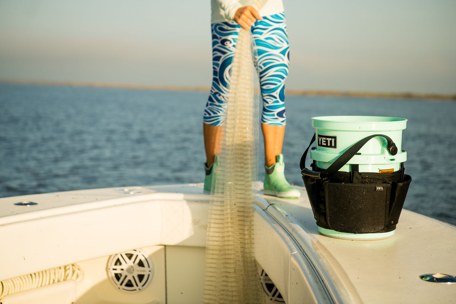 Eine Person auf einem Boot in blau gemusterten Leggings und grünen Schuhen hält ein Fischernetz; eine schwarze YETI® Gürteltasche für Loadout 19 Liter Eimer sitzt auf dem Deck nahe dem Wasser.