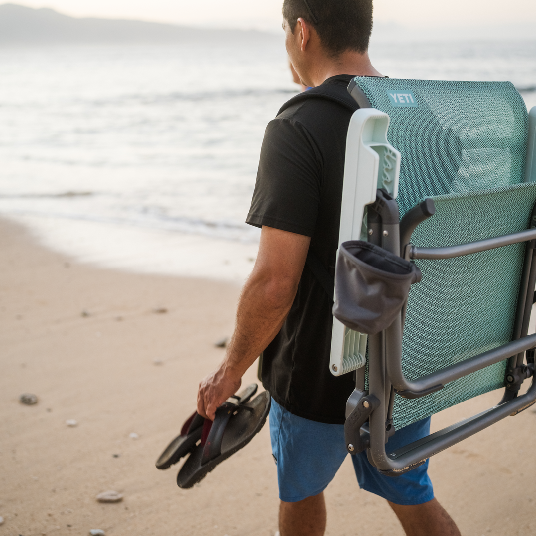 Bei Sonnenuntergang schlendert eine Person mit einem gefalteten YETI® Hondo Beach Chair - Navy von YETI auf dem Rücken und Flip-Flops in der Hand einen Sandstrand entlang, während im Hintergrund die Wellen des Ozeans und die ferne Küste zu sehen sind.