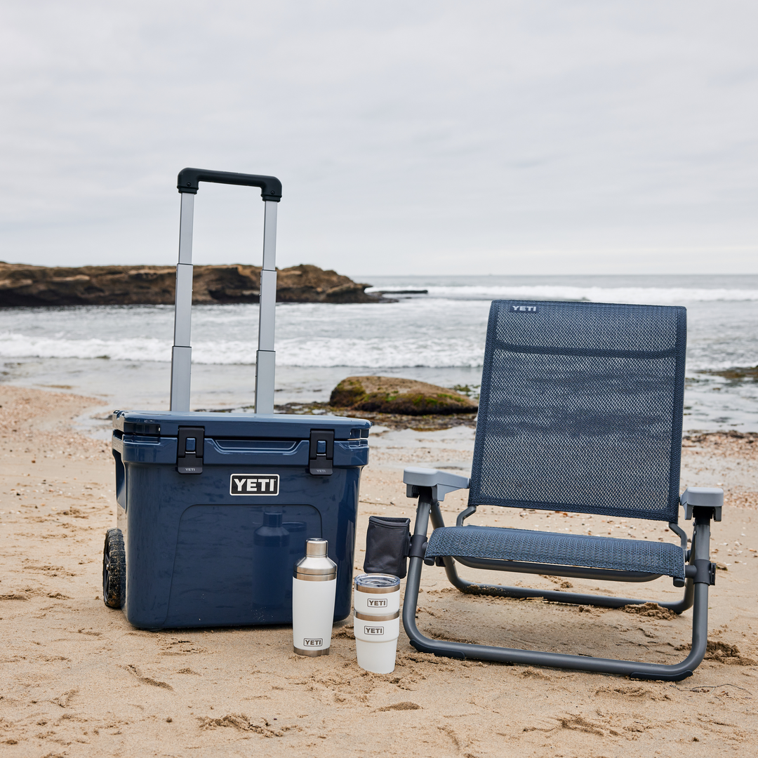 YETI® Hondo Beach Chair - Navy, eine rollende marineblaue Kühlbox, ein weißer Becher und ein isolierter Becher am Sandstrand mit Meereswellen und felsiger Küste im Hintergrund.