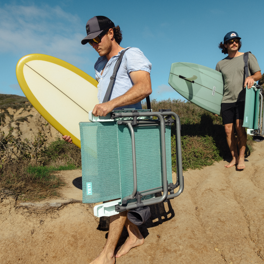 Zwei Personen laufen barfuß auf einem Sandweg mit Surfbrettern und einem YETI® Hondo Beach Chair - Navy in Richtung Strand an einem sonnigen Tag mit Büschen und blauem Himmel im Hintergrund.