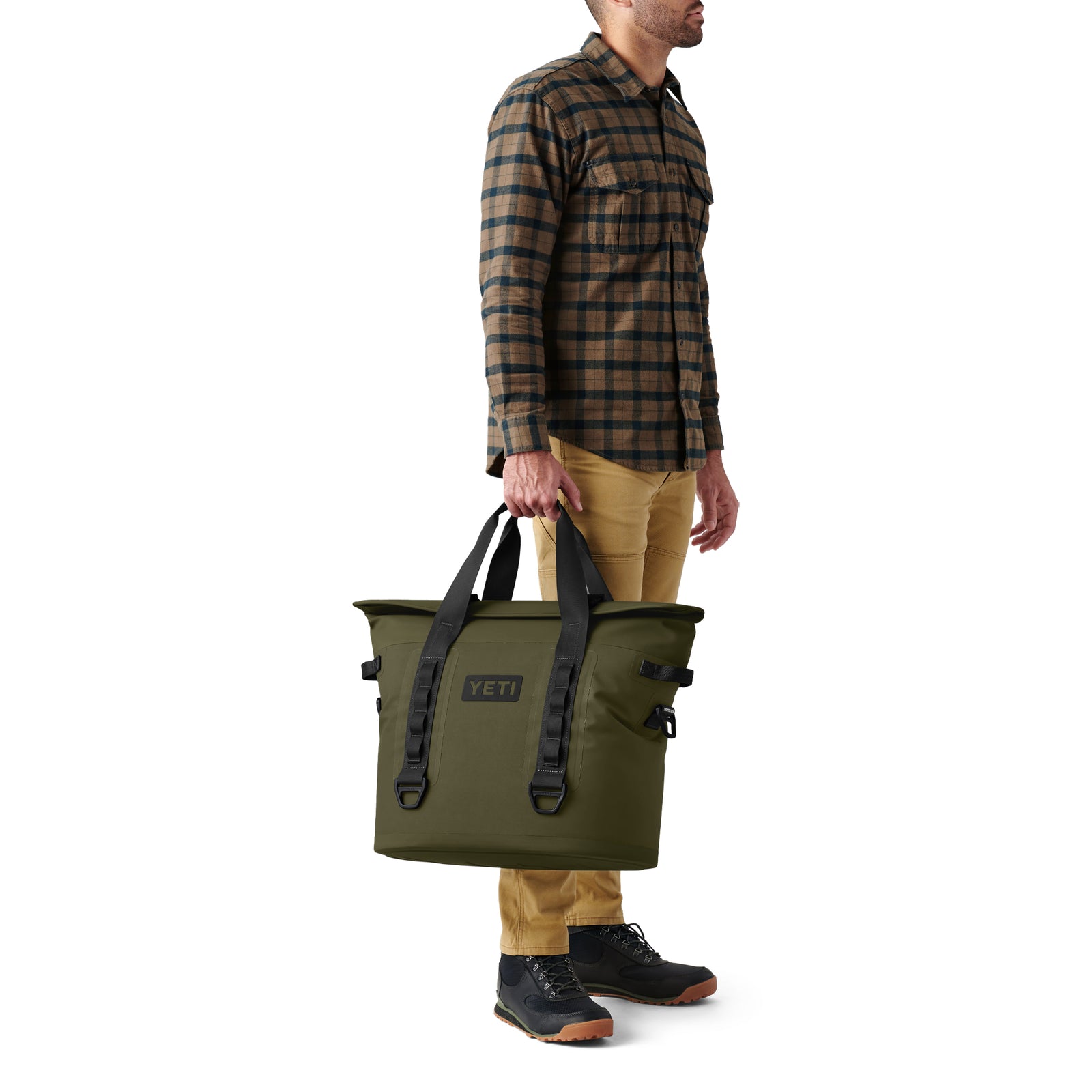 Ein Mann in einem braun karierten Hemd, einer hellbraunen Hose und schwarzen Schuhen hält eine YETI® Hopper M30 Soft Cooler - Olive an den Griffen vor einem weißen Hintergrund.