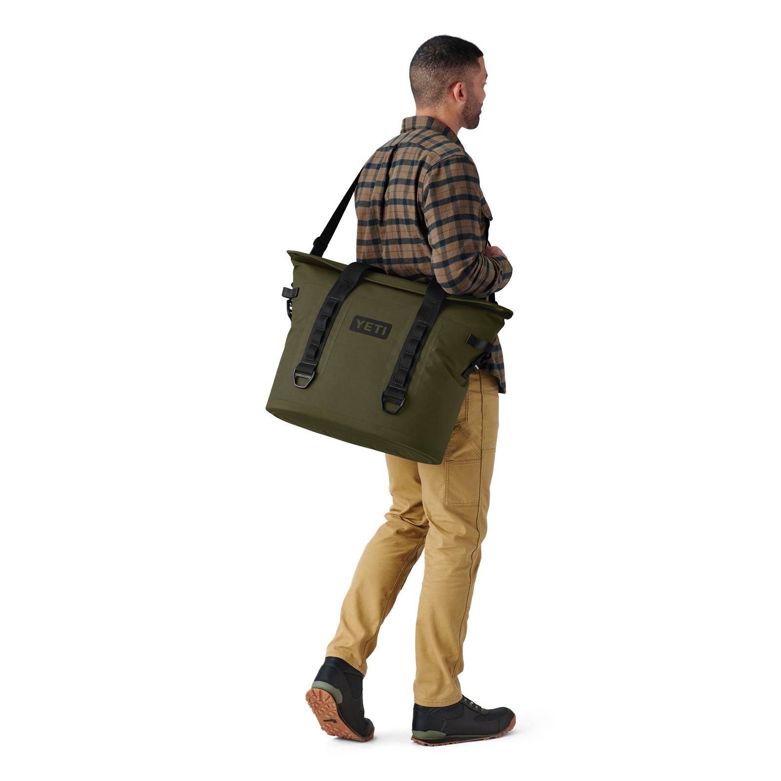 Ein Mann in einem karierten Hemd, einer hellbraunen Hose und schwarzen Schuhen trägt eine YETI® Hopper M30 Soft Cooler - Olive von YETI und geht vor einem weißen Hintergrund von der Kamera weg.