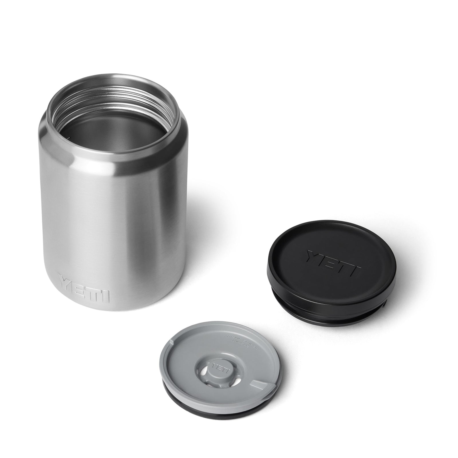 Ein YETI® Isolierter Essensbehälter Rambler 24 oz aus Edelstahl steht auf einer weißen Fläche mit zwei schwarzen und grauen Deckeln in der Nähe, von denen einer das YETI-Logo zeigt. Der geöffnete Behälter zeigt seinen leeren Innenraum und die doppelwandige Vakuumisolierung für beste Temperaturhaltung.