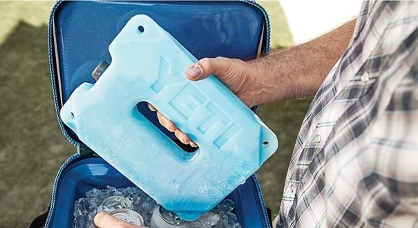 Eine Hand legt einen blauen YETI® Kühlakku Ice Block 2 lb in eine offene isolierte Kühltasche, die mit Eis und einem Dosengetränk gefüllt ist, und unterstreicht damit das robuste Design von YETI für lang anhaltende Kühlung bei jedem Abenteuer.