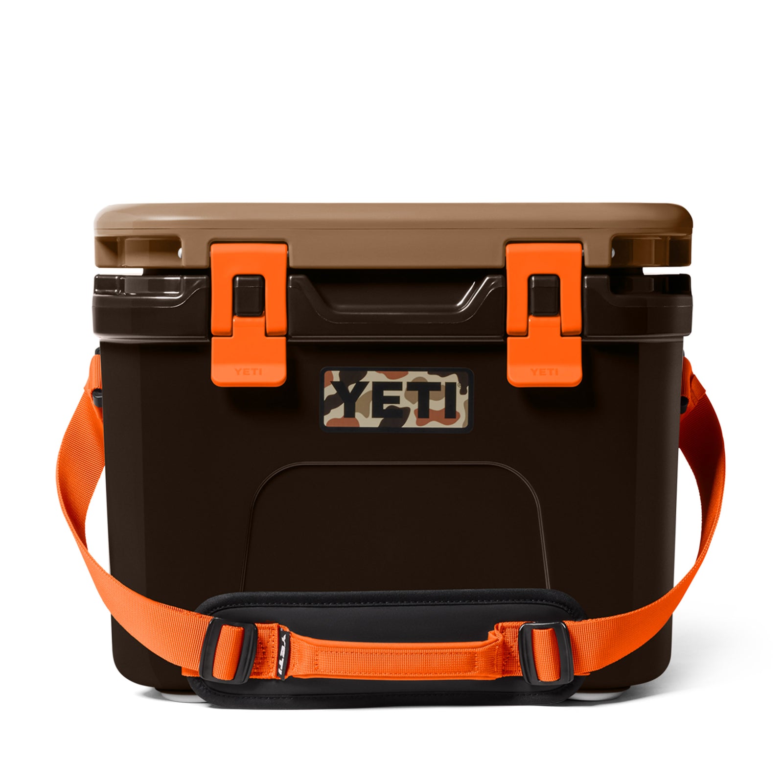 Die YETI® Kühlbox Roadie 15 - Wetlands Camo hat einen braunen Körper, einen hellbraunen Deckel, orangefarbene Verschlüsse, ein YETI-Logo in Tarnfarben und einen orangefarbenen Schultergurt mit schwarzer Polsterung auf weißem Hintergrund.