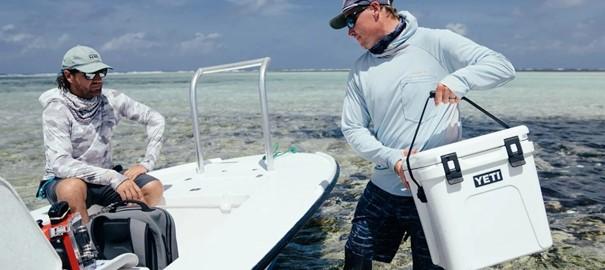 Zwei Männer in Sonnenschutzkleidung befinden sich auf einem kleinen Boot im flachen, klaren Wasser. Der eine hält eine YETI® Kühlbox Roadie 24 - Navy in der Hand, während der andere mit einer grauen Tasche unter einem teilweise bewölkten Himmel sitzt.