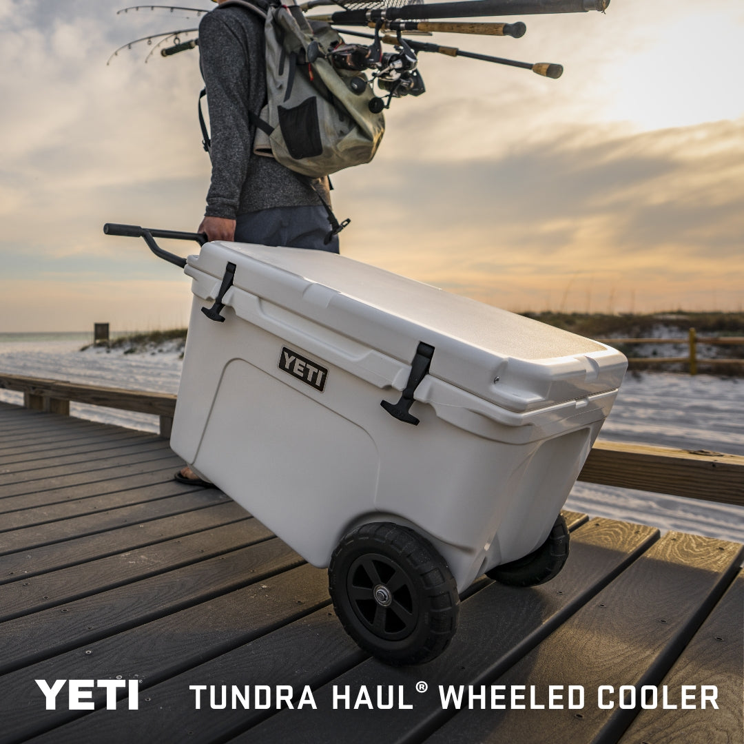 Eine Person zieht eine weiße YETI® Kühlbox Tundra Haul bei Sonnenuntergang an einer Strandpromenade entlang und trägt Angelausrüstung und Ruten. Text: YETI® Kühlbox Tundra Haul - Weiß. Marke: YETI.