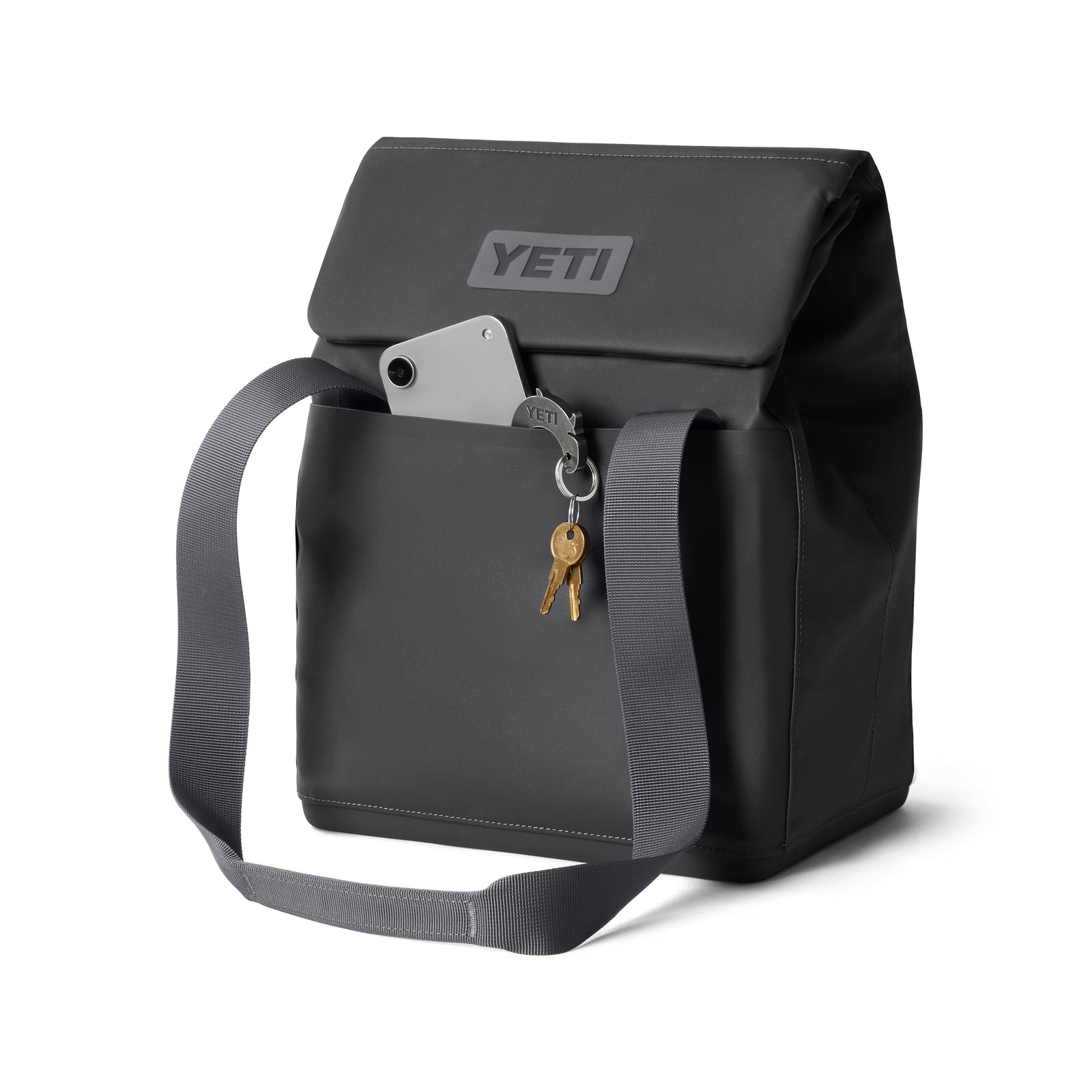Eine anthrazitfarbene YETI® Kühltasche Daytrip Lunch Bag 14L mit verstellbarem, grauem Gurt, Klappenverschluss, sichtbarem YETI-Logo, Vordertasche für ein Smartphone und einem Schlüsselanhänger mit zwei Schlüsseln. Mit Coldcell Flex Isolierung.