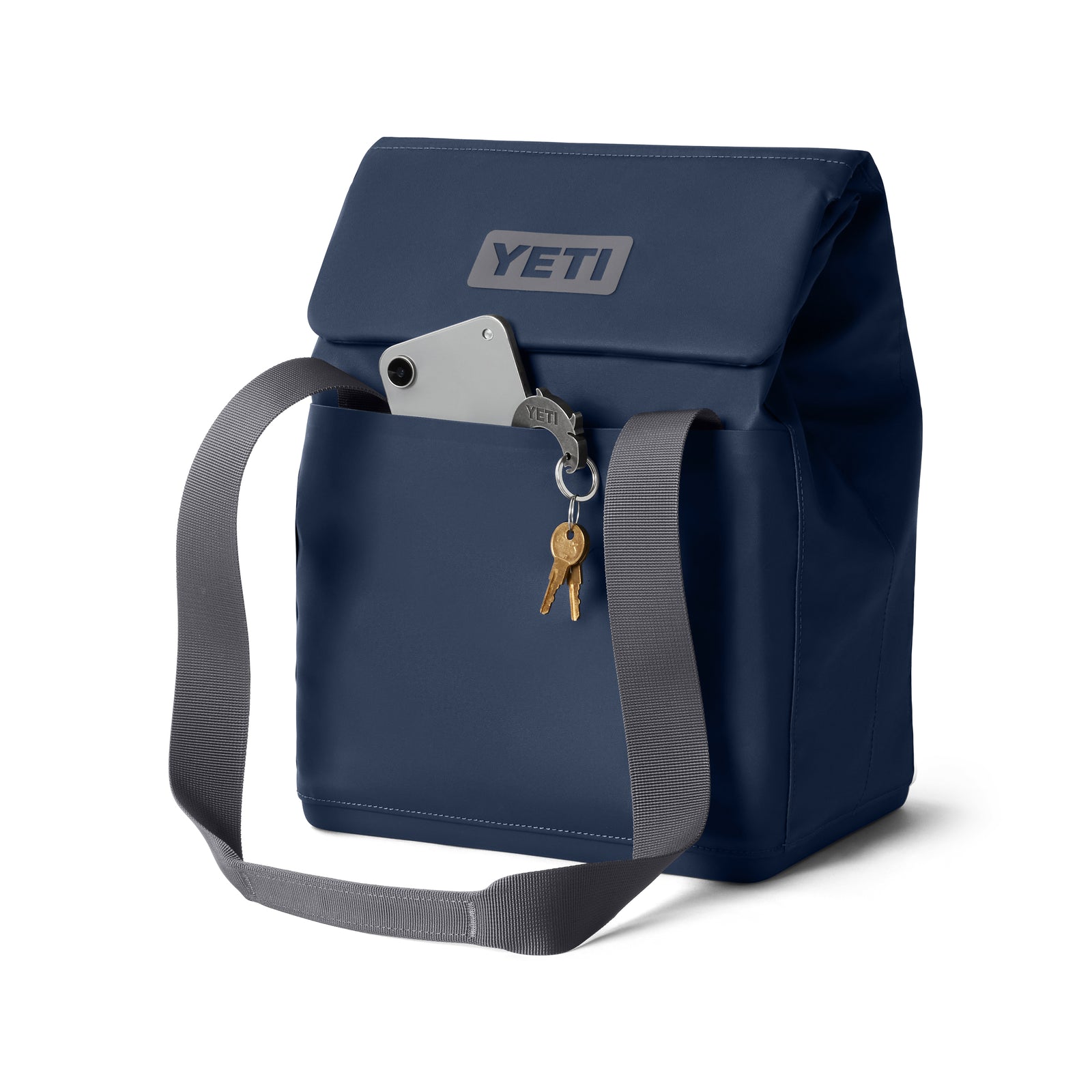 Eine YETI® Kühltasche Daytrip Lunch Bag 14L in Navy mit Coldcell Flex-Isolierung, grauem Schultergurt, Fronttasche mit Smartphone und Schlüsselanhänger mit zwei Schlüsseln vor weißem Hintergrund.