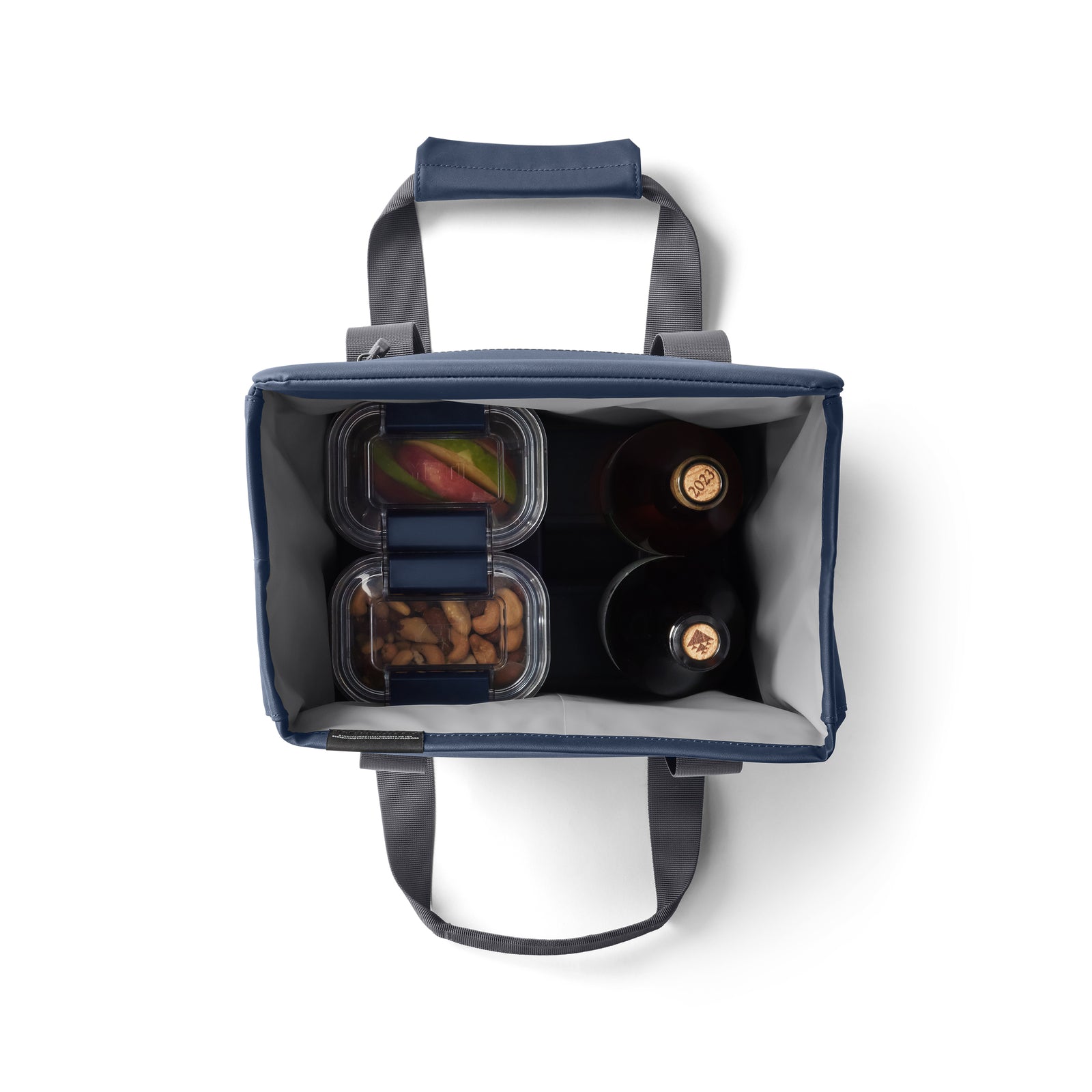 Draufsicht auf die YETI® Kühltasche Daytrip Lunch Bag 14L - Navy mit grauen Griffen, zeigt zwei Flaschen, einen durchsichtigen Behälter mit geschnittenem Obst und einen mit Mandeln. Die Coldcell Flex Isolierung von YETI hält den Inhalt frisch.