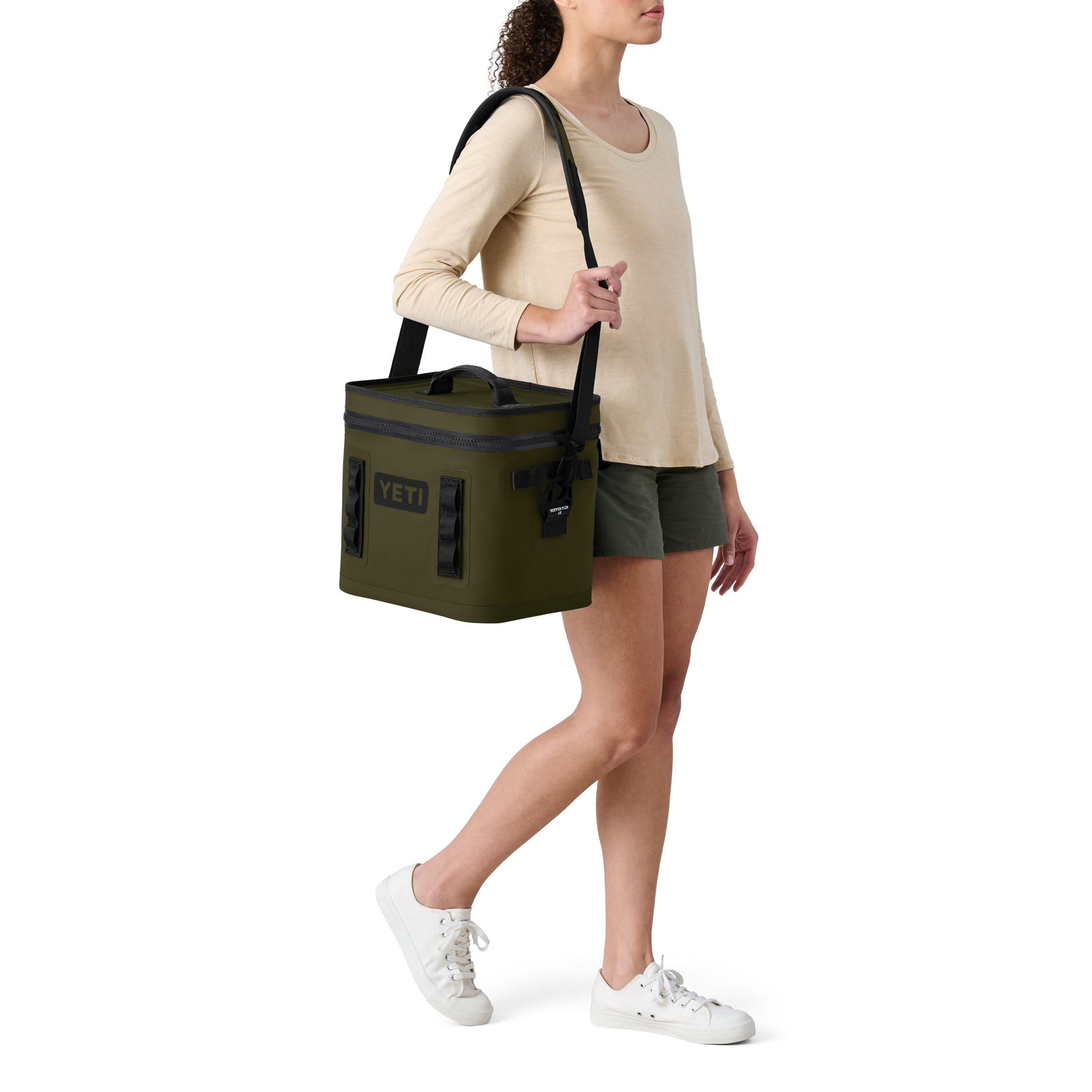 Eine Frau trägt die YETI® Kühltasche Hopper Flip® 12 in Olive mit einem Schulterriemen. Sie trägt ein beigefarbenes langärmeliges Hemd, grüne Shorts und weiße Turnschuhe und steht vor einem schlichten weißen Hintergrund.