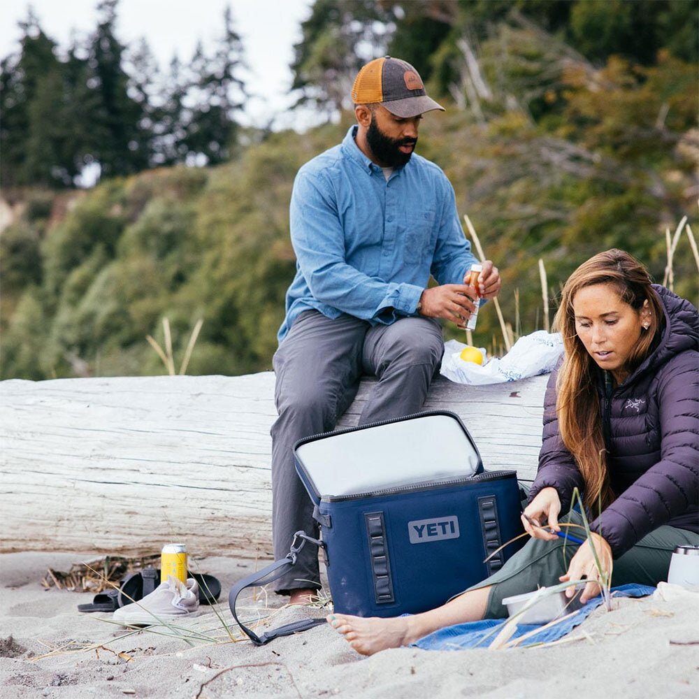 Zwei Personen entspannen sich an einem Sandstrand mit der YETI® Kühltasche Hopper Flip® 18 in Navy. Ein Mann sitzt auf einem Treibholzstamm und hält einen Drink in der Hand, während eine Frau neben der YETI Kühltasche sitzt.