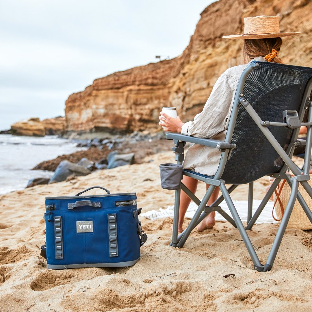 Eine Person mit Strohhut entspannt sich an einem Sandstrand bei felsigen Klippen und hält eine Tasse in der Hand. In der Nähe ruht die YETI® Kühltasche Hopper Flip® 18 - Navy von YETI auf dem Sand mit dem Meer und dem bewölkten Himmel im Hintergrund.