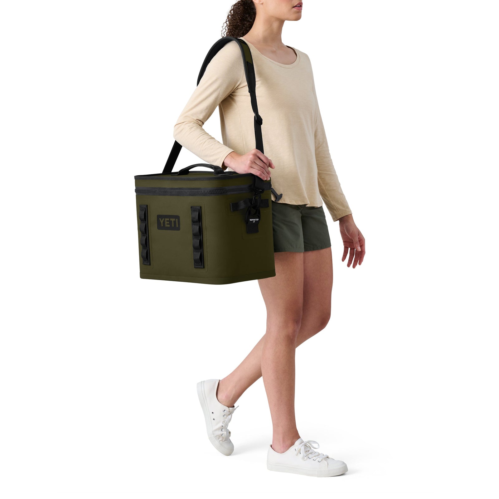 Eine Person in einem beigen langärmeligen Hemd, grünen Shorts und weißen Turnschuhen trägt die YETI® Kühltasche Hopper Flip® 18 - Olive Soft Cooler mit Schulterriemen von YETI vor einem weißen Hintergrund.