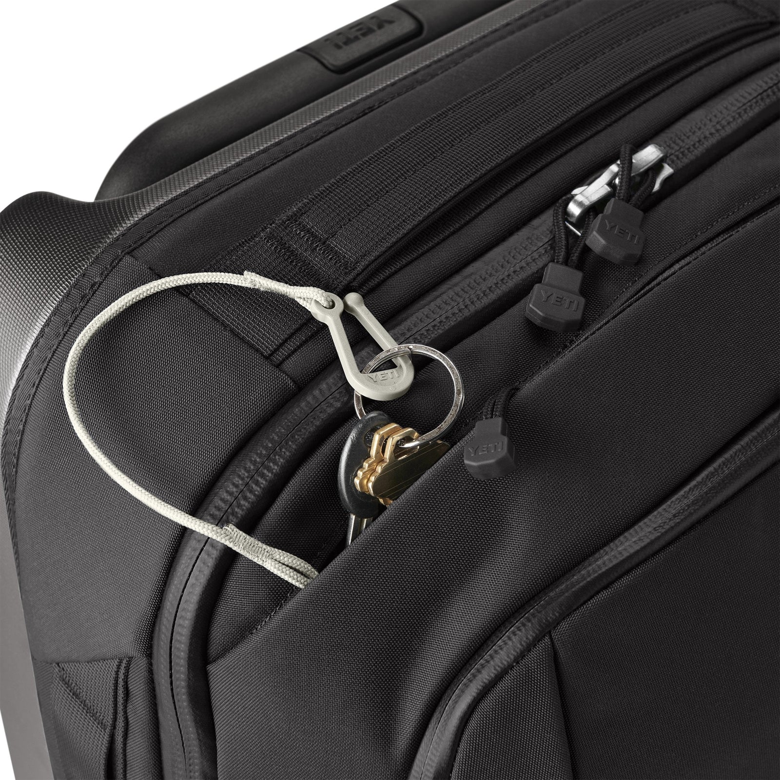 Eine Nahaufnahme einer schwarzen Koffertasche aus TuffSkin-Nylon mit einem Schlüsselring an einem weißen Sicherheitsclip neben den Reißverschlüssen - ideal für organisierte Reisende, die den YETI® Koffer Crossroads® 22 - Black von YETI verwenden.