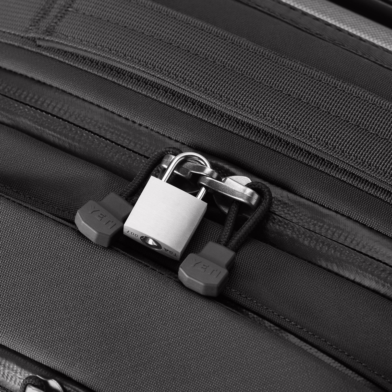 Ein silbernes Vorhängeschloss sichert die Reißverschlüsse des schwarzen YETI® Koffer Crossroads® 22, der aus TuffSkin-Nylon gefertigt ist, und zeigt, dass die Marke auf Sicherheit und Langlebigkeit bei Reiseausrüstung setzt.