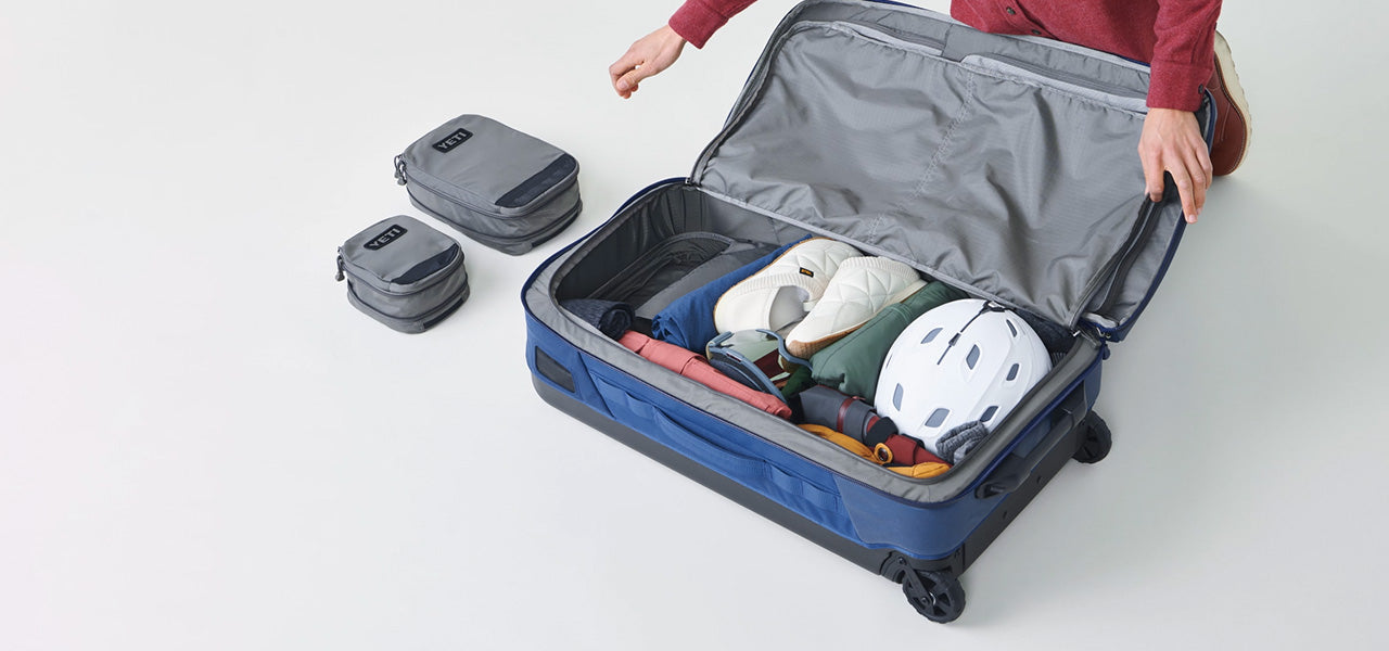 Eine Person kniet neben einem geöffneten YETI® Koffer Crossroads® 22 - Black aus TuffSkin Nylon, gefüllt mit Kleidung, Schuhen und einem weißen Helm. Zwei graue Packwürfel stehen neben dem Koffer auf einer weißen Fläche.