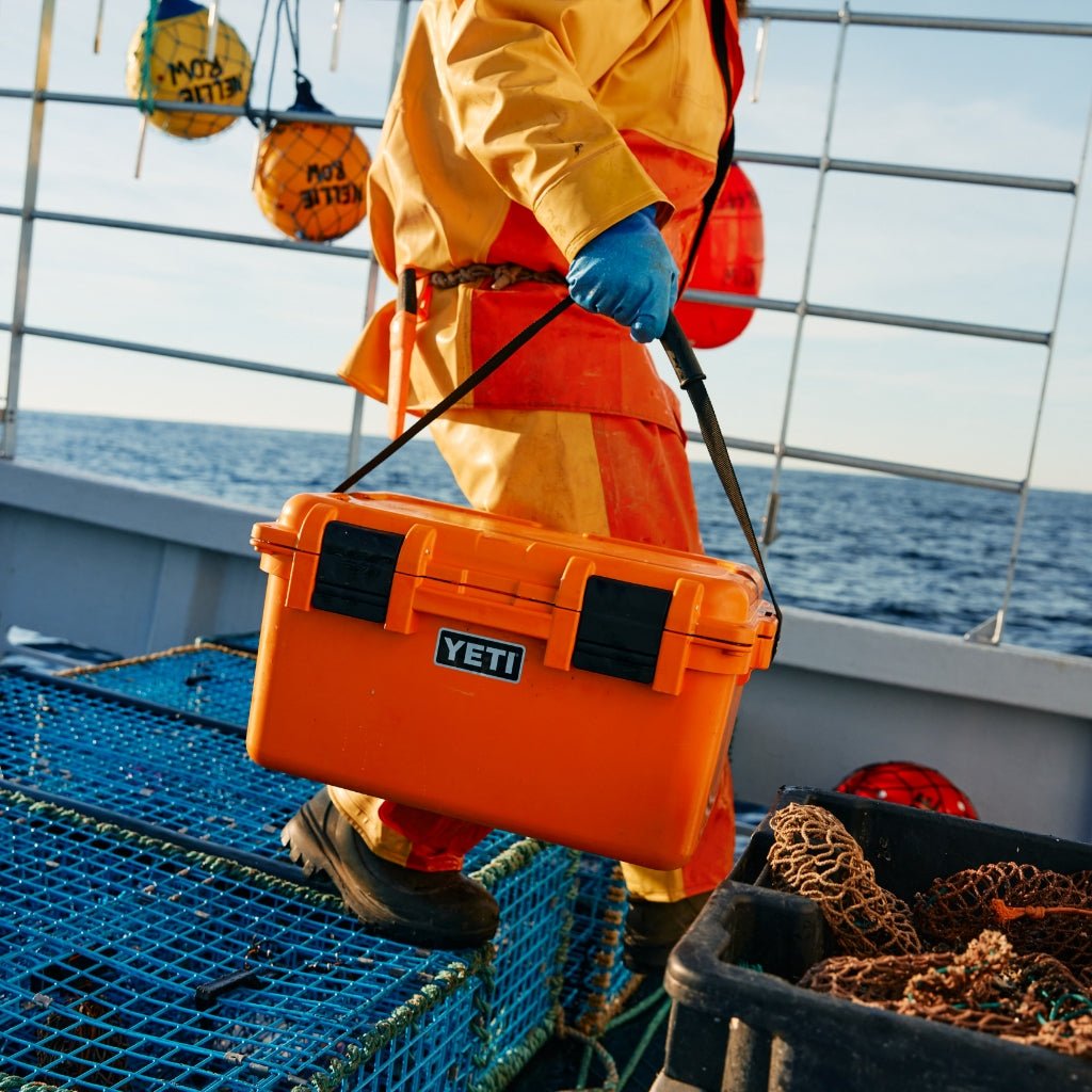 Eine Person in orangefarbener wasserdichter Kleidung und blauen Handschuhen trägt die YETI® LoadOut GoBox 30 - King Crab Orange auf einem Fischerboot mit Hummerfallen und dem Meer im Hintergrund.
