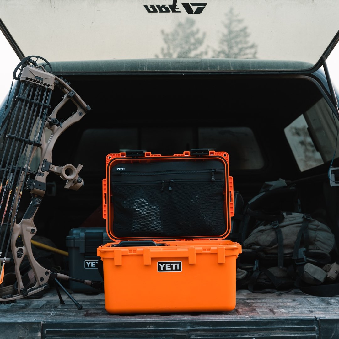 Eine offene YETI® LoadOut GoBox 30 in King Crab Orange liegt auf einer LKW-Heckklappe neben einem Bogen, einem Rucksack und anderer Ausrüstung, mit Bäumen im Hintergrund.